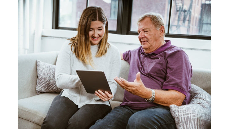 A daughter sitting on a couch with her dad, showing him the Sold Out Products collection on a tablet on Doms Mattress Store website, and explaining how the store’s products have evolved over time