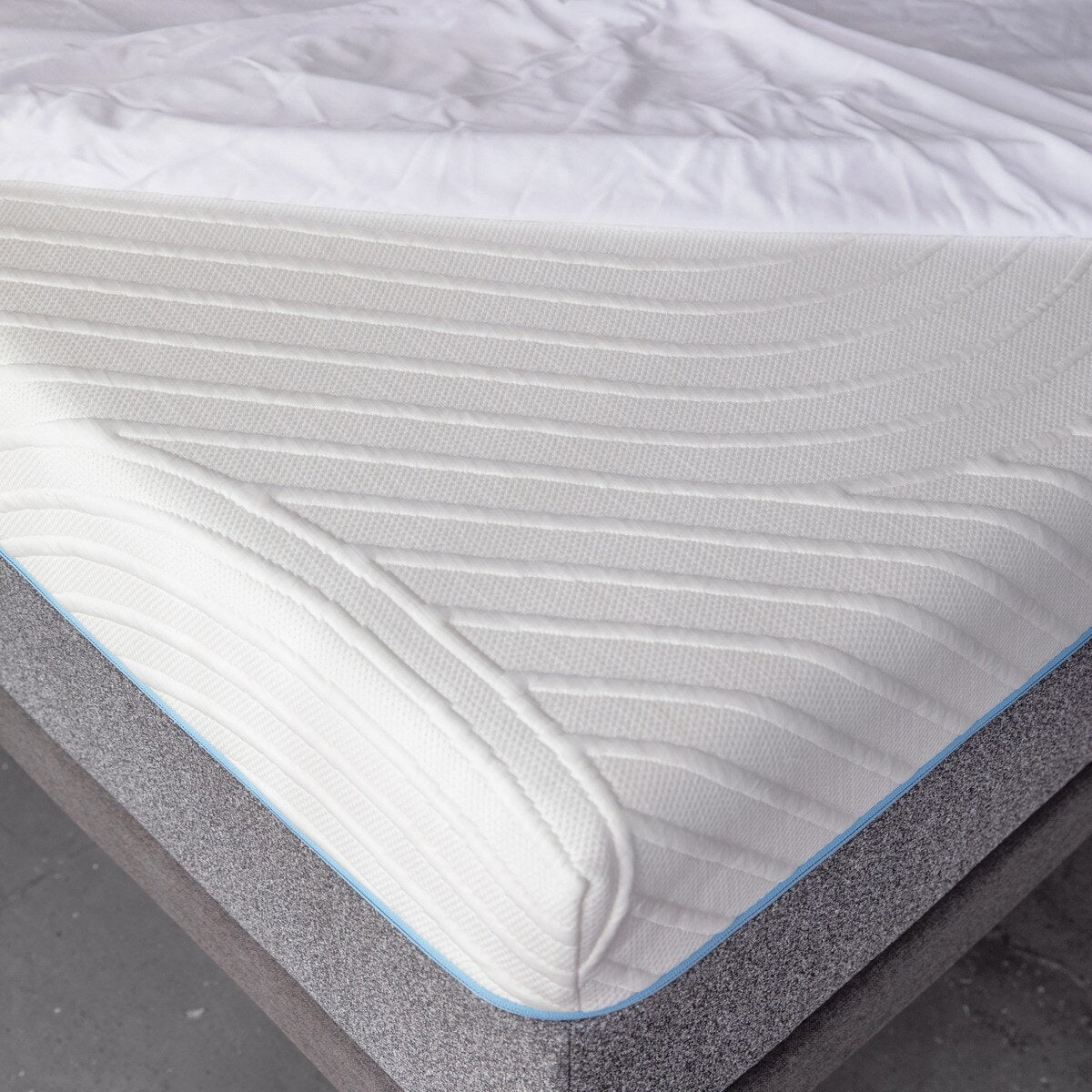 Close up corner view of MLILY Polyester Knit Fabric Mattress Protector on mattress, placed on platform bed frame, mattress protector is only halfway made over the mattress to show that it covers the mattress like a fitted sheet, concrete floor.