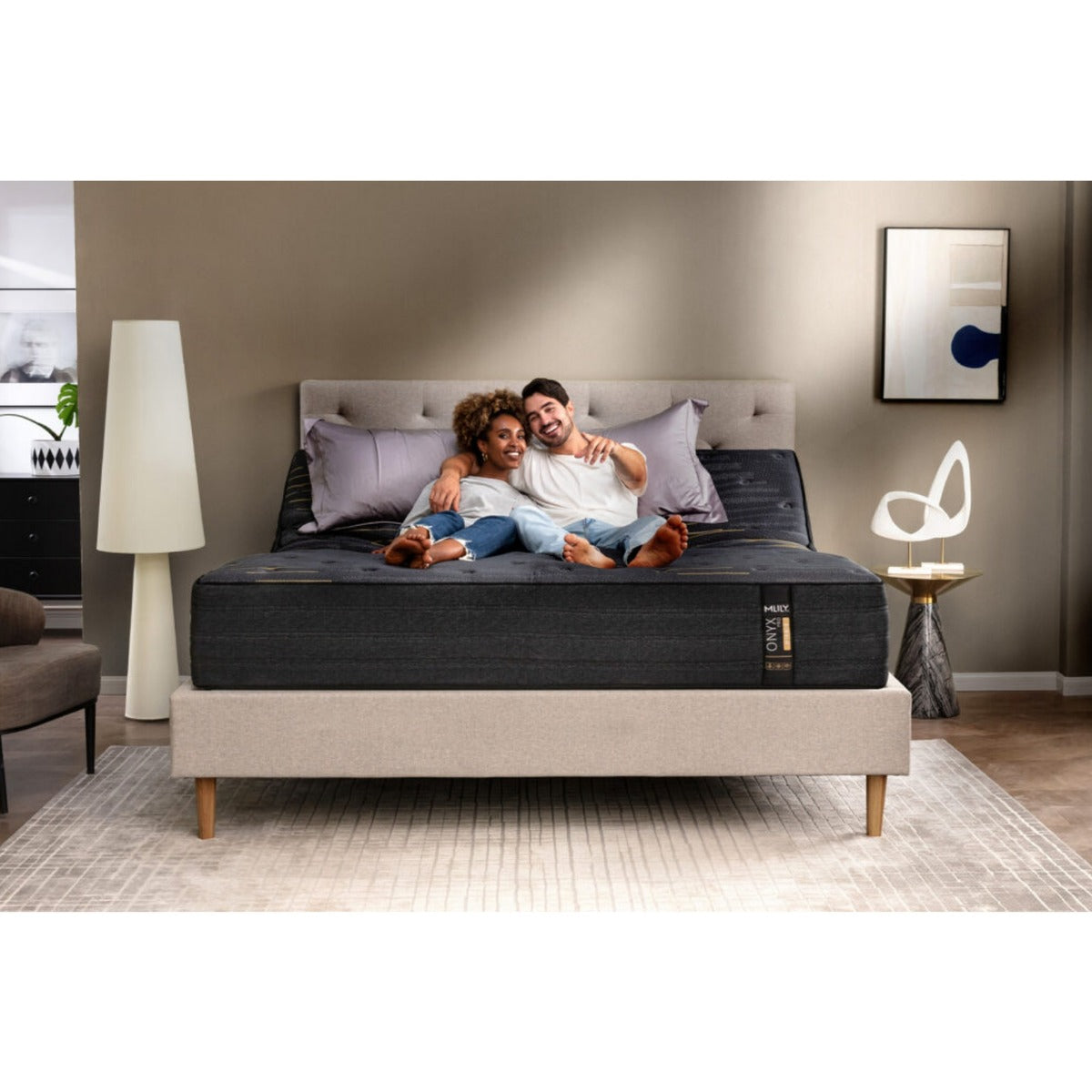 Front view of the MLILY ONYX Pro Hybrid Medium 13" Mattress on a platform base, with the head of the mattress raised, pillows at the head, a man and woman lying on the bed holding each other while watching TV, in a decorated bedroom.