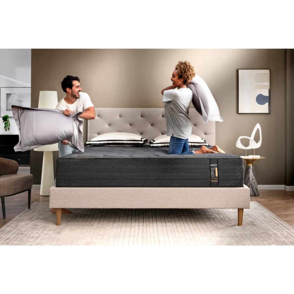 Front view of the MLILY ONYX Pro Hybrid Medium 13-inch Mattress on a platform bed frame, man and woman having a playful pillow fight to show the mattress support, with pillows at the head of the bed, decorated bedroom.