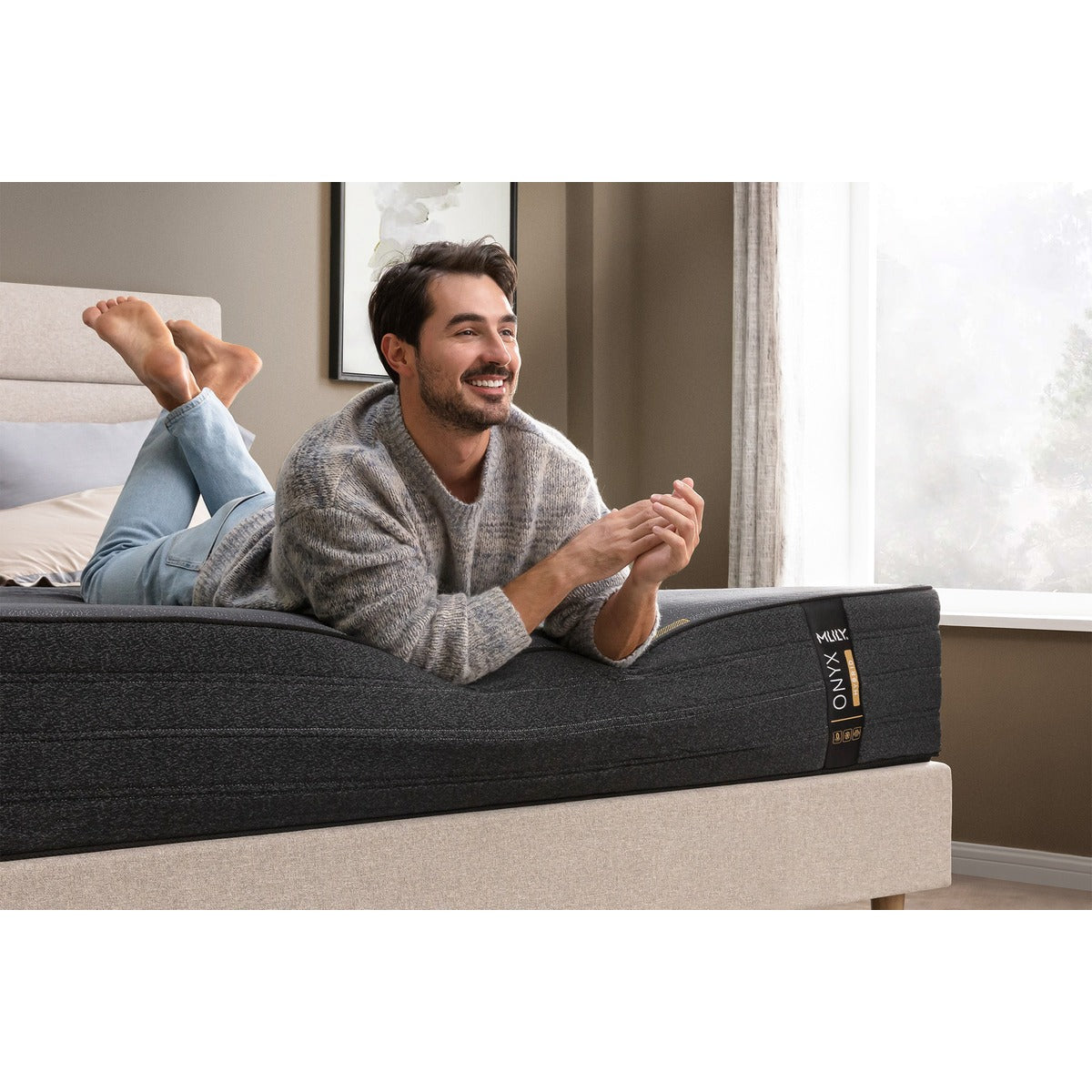 MLILY ONYX Hybrid Firm 12-inch Mattress, on a platform bed frame, with a man laying on his stomach at the foot of the mattress, demonstrating edge support, inside a decorated bedroom with pillows at the head.