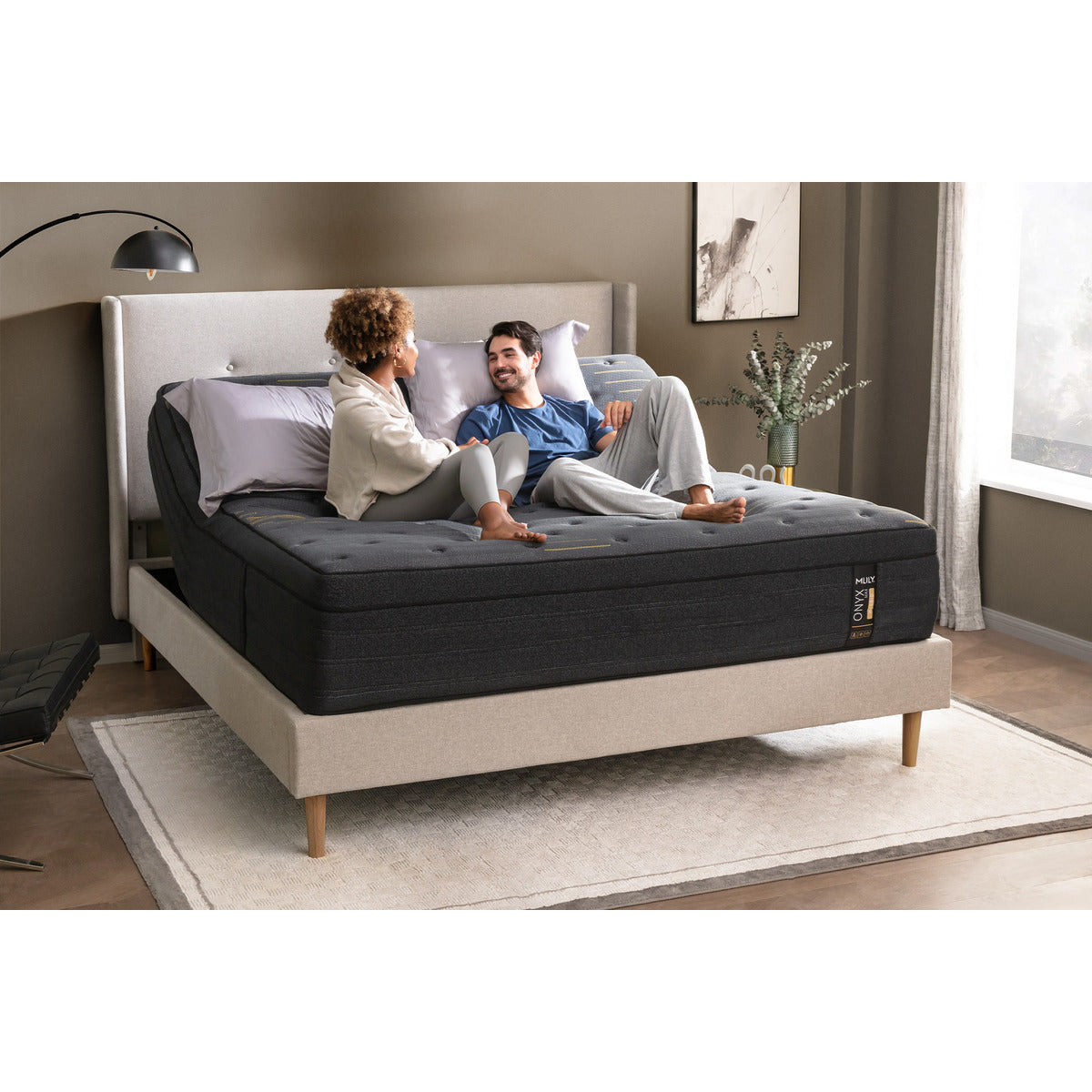 MLILY ONYX Max Hybrid Plush 14-inch Mattress corner view with side and front visible, adjustable base raised at the head, pillows arranged, a couple relaxing on the bed, surrounded by a platform bed frame in a decorated bedroom.