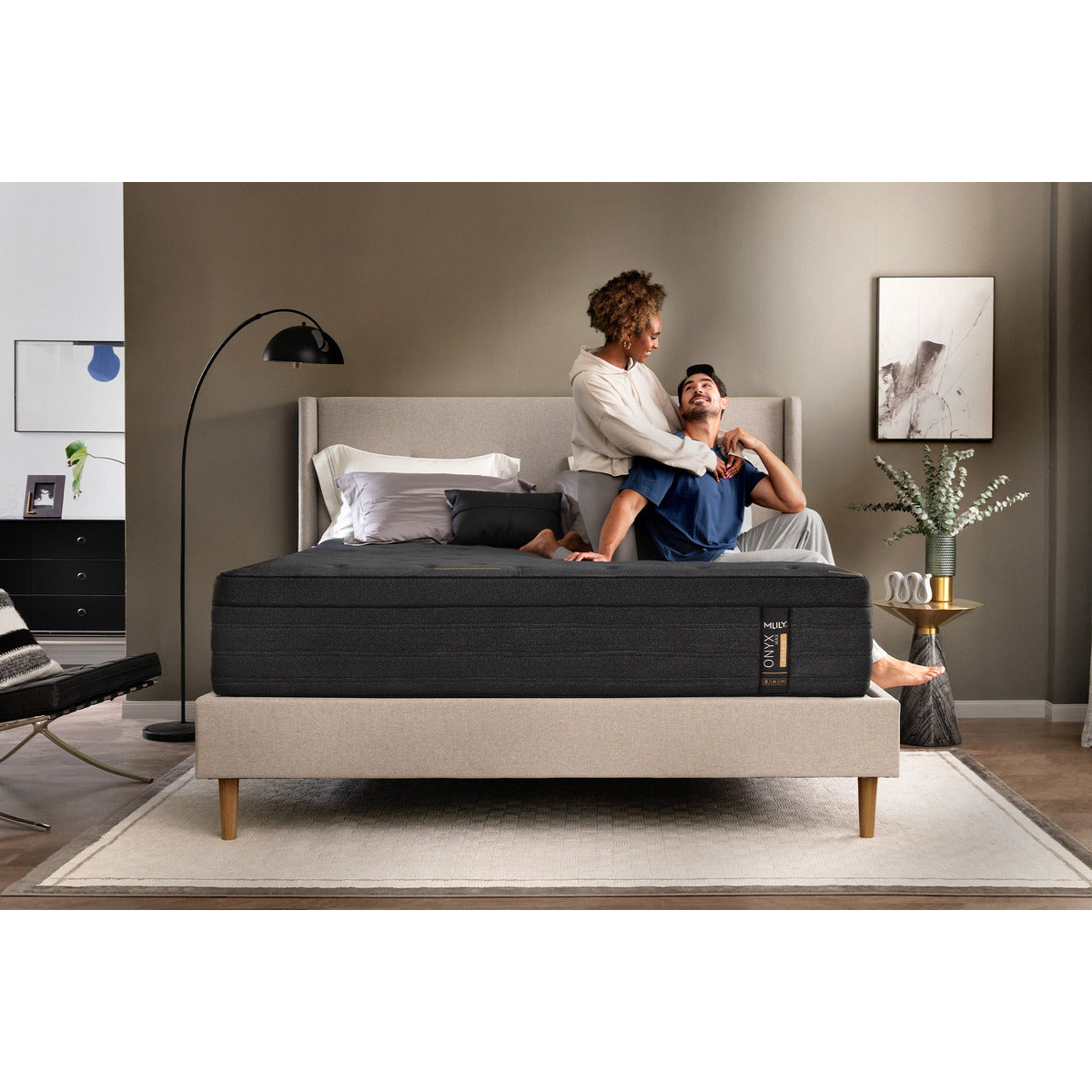 MLILY ONYX Max Hybrid Plush 14-inch Mattress on platform bed frame, facing forward, man and woman sitting on one side of the bed, woman with arms wrapped around man, a few pillows at the head of the mattress, decorated bedroom.