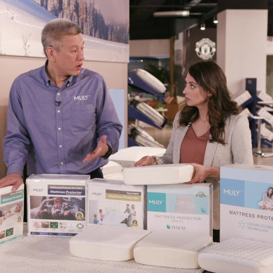 A salesman explains the MLILY Polyester Knit Fabric Mattress Protector to a woman, highlighting its breathability, dust mite and mildew resistance, 100% waterproof features, and fitted sheet-like design.
