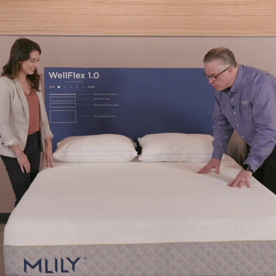 A salesman explains the MLILY WellFlex 1.0 Memory Foam Firm 12" Mattress to a woman, emphasizing its firm support, spine alignment, cooling ice fabric cover, and benefits for stomach sleepers.