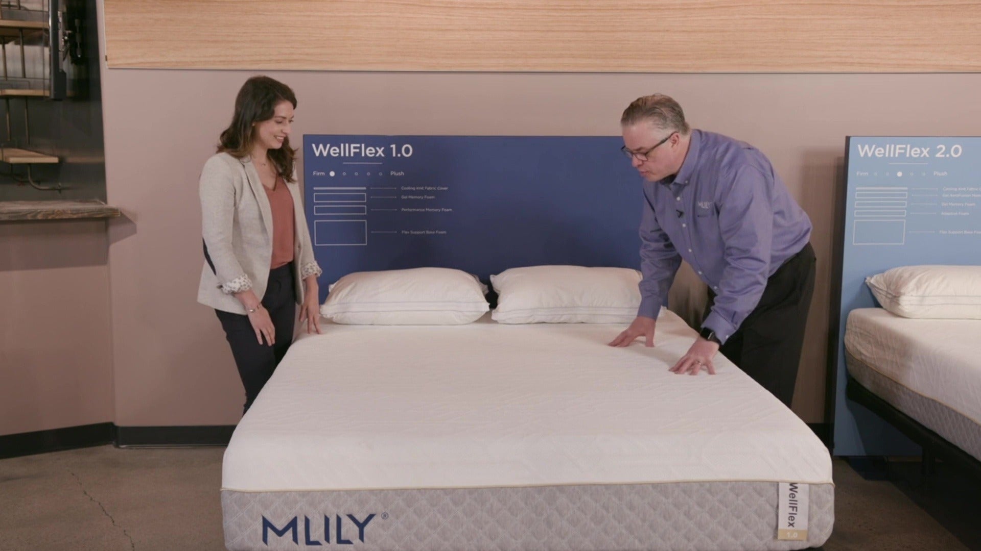 A salesman explains the MLILY WellFlex 1.0 Memory Foam Firm 12" Mattress to a woman, emphasizing its firm support, spine alignment, cooling ice fabric cover, and benefits for stomach sleepers.