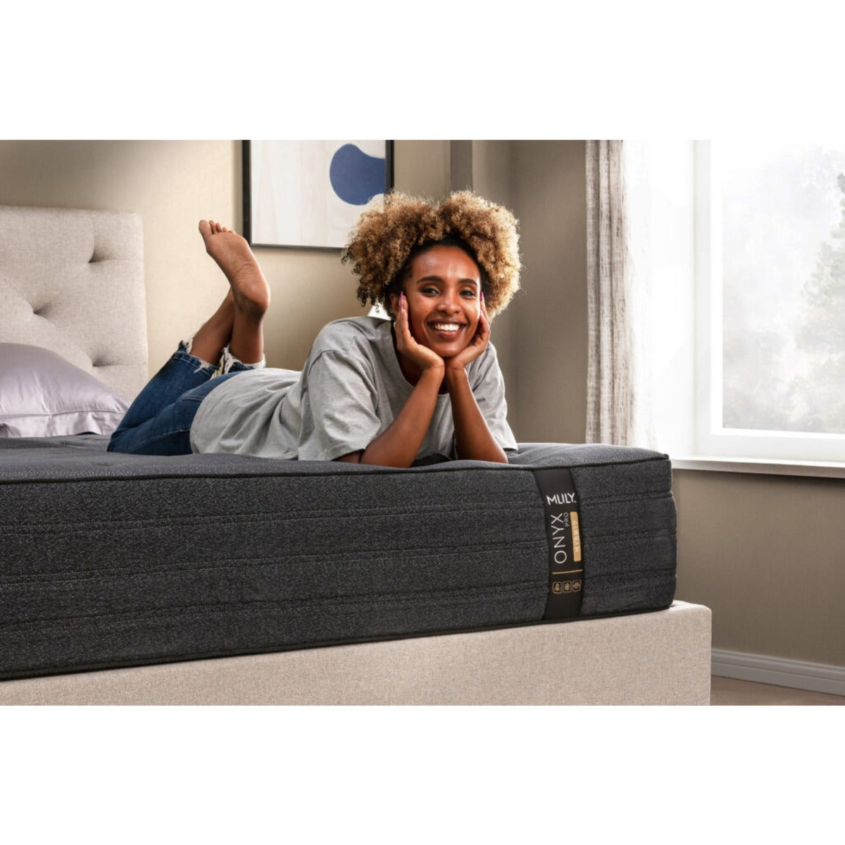 Side view of the MLILY ONYX Pro Hybrid Medium 13" Mattress on a platform bed frame, pillow at the head, woman laying on her stomach at the foot of the mattress while sitting up, demonstrating the support of the bed, decorated bedroom.