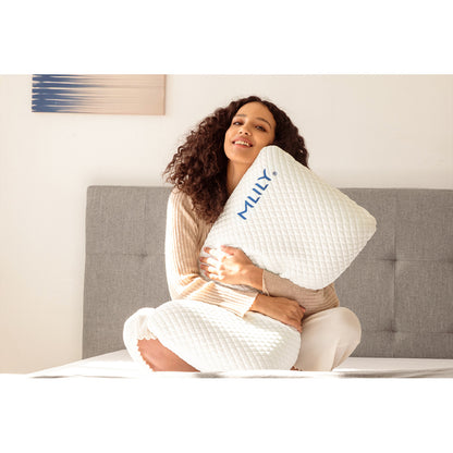 Woman sitting on a mattress hugging the MLILY Serenity Cool Pillow, demonstrating the comfort of the Ventilated gel-infused AeroFusion Memory Foam core.