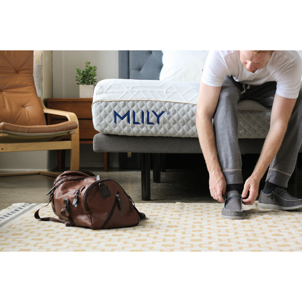 Zoomed-in front corner view of the foot of the MLILY WellFlex 1.0 Memory Foam Firm 12" Mattress, with a man sitting at the edge putting on his work shoes, demonstrating the supportiveness of the mattress edge, and a travel bag placed nearby.